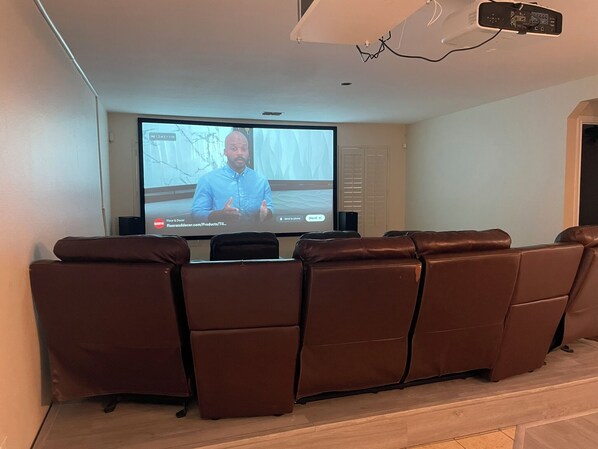 A home theater setup with a large flat-screen TV mounted on the wall, leather reclining chairs, and a ceiling-mounted projector. - Charming 4BR Getaway with 130” Theater, WiFi & AC in San Antonio (San Antonio)