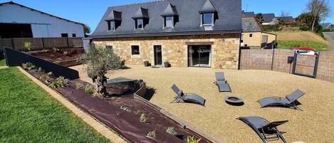 A stone building with a sloped roof and a patio area with lounge chairs and a fire pit.
