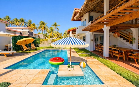 A swimming pool with a floating watermelon, a striped umbrella, and a beach ball.