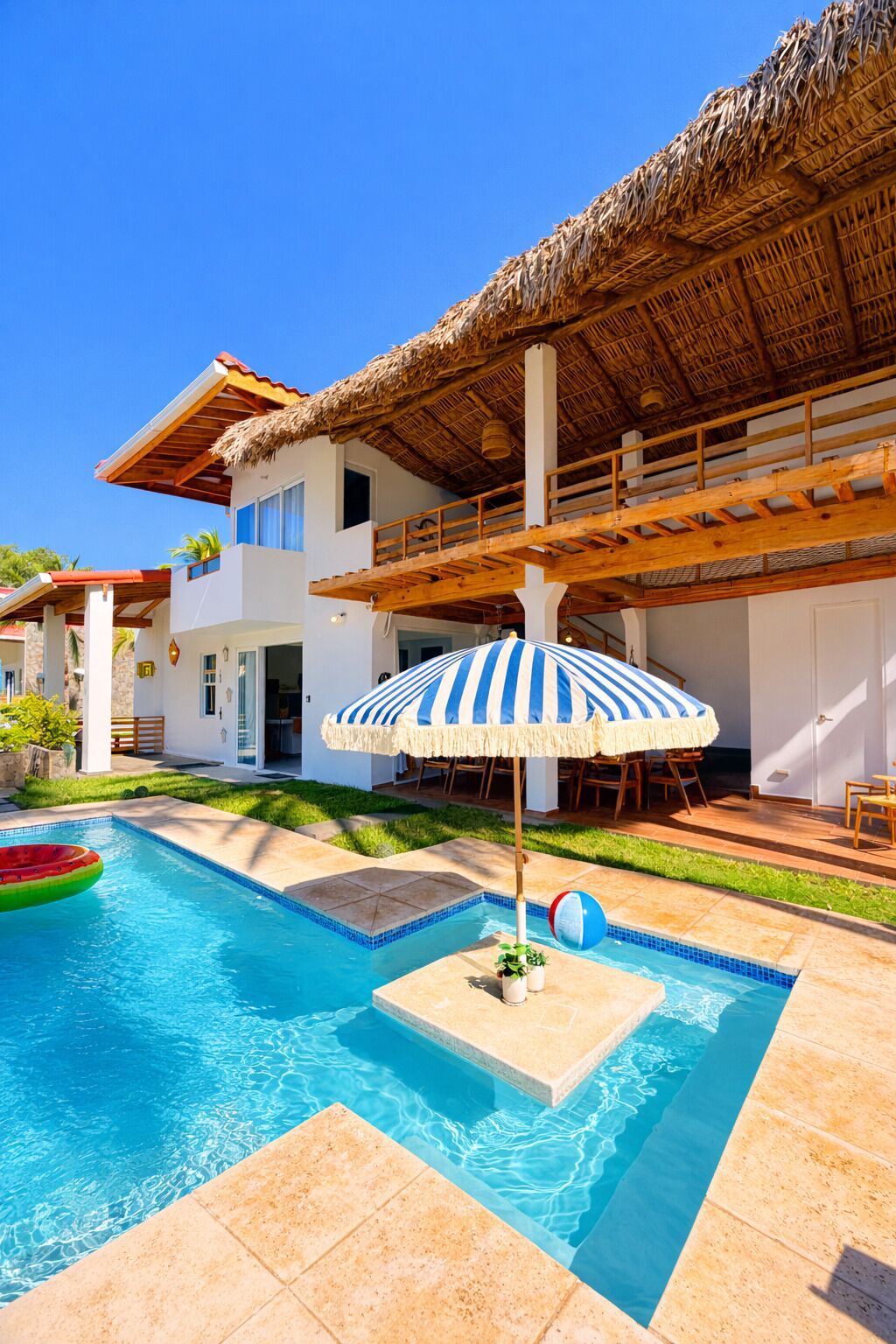 A modern house with a pool, a thatched roof, and a blue-striped umbrella.