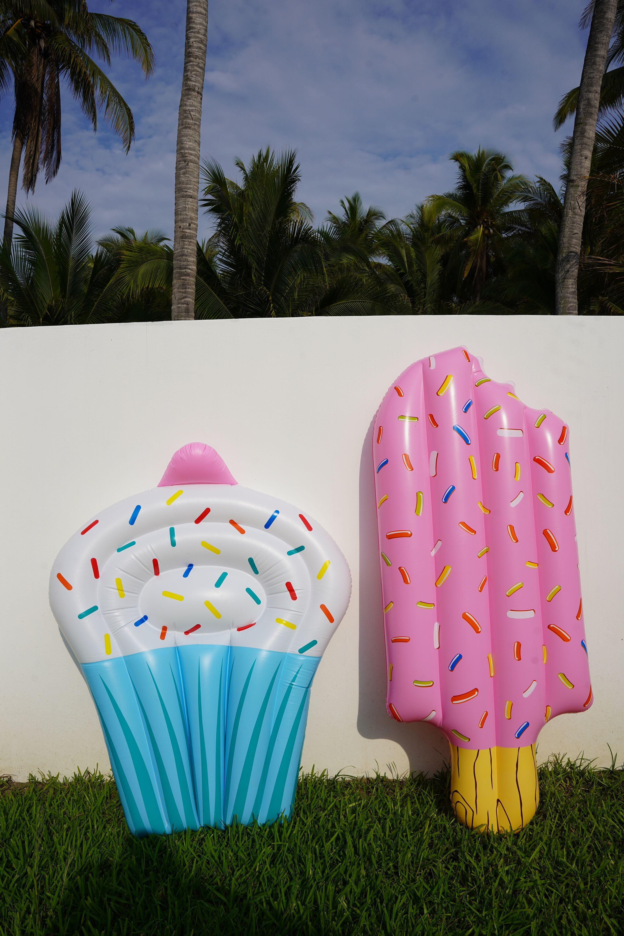 Dos flotadores inflables con forma de cupcake y paleta sobre un césped, con palmeras al fondo.