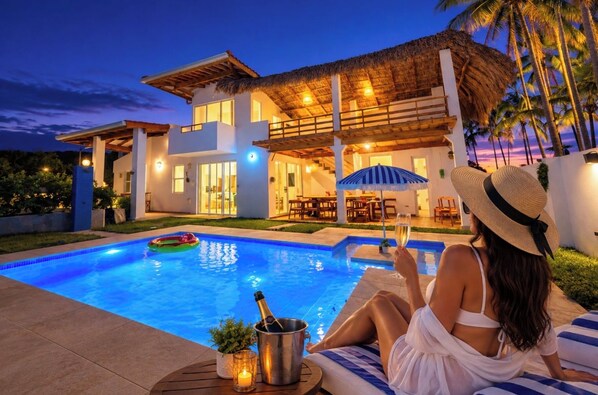 Una casa moderna con piscina, mobiliario exterior y una estructura de techo de paja.