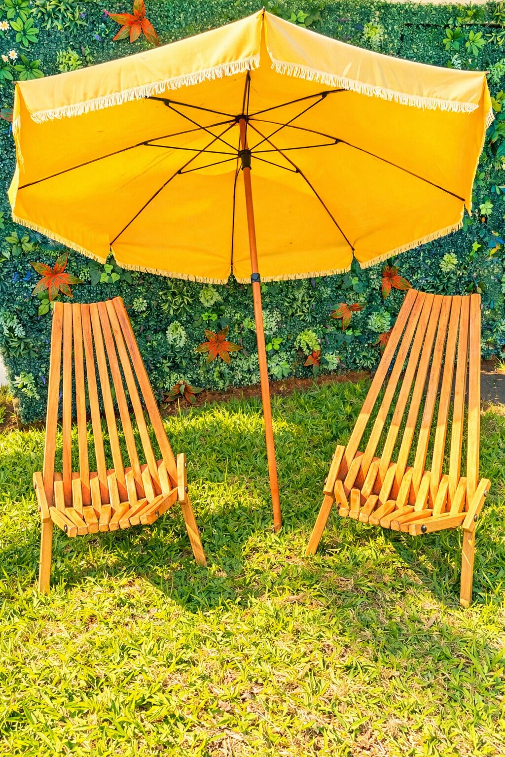 Two wooden chairs with a yellow umbrella between them, set on grass with a hedge in the background.
