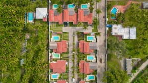 Vista aérea de un resort con varias construcciones, cada una con techo rojo y piscina. El resort está junto a una playa con olas rompiendo en la orilla.