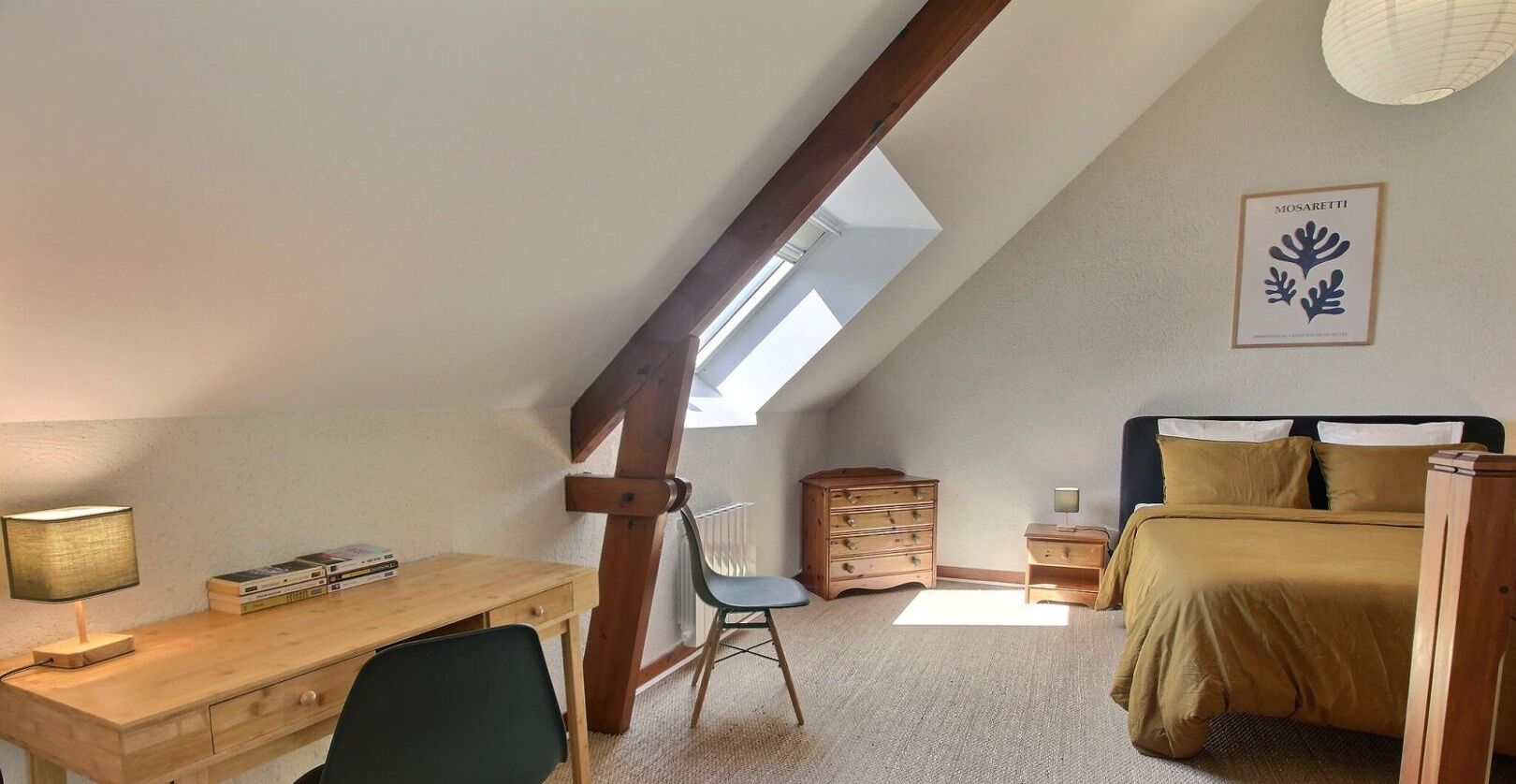 Una camera da letto con soffitto spiovente, una scrivania con lampada, una sedia, un letto con testiera, una cassettiera e un quadro incorniciato appeso al muro.
