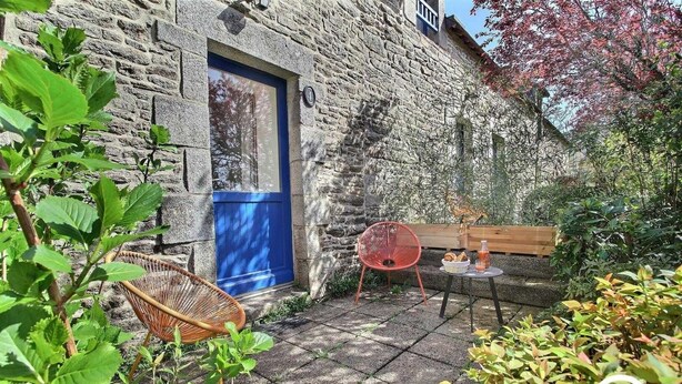A stone building with a blue door, a wicker chair, a red chair, and a small table with a drink and snacks. - La Ferme rouge, Le Pin (PLUVIGNER)