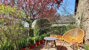Un patio con una mesa redonda y una silla de mimbre, rodeado de plantas en macetas y un árbol de hojas rojas.