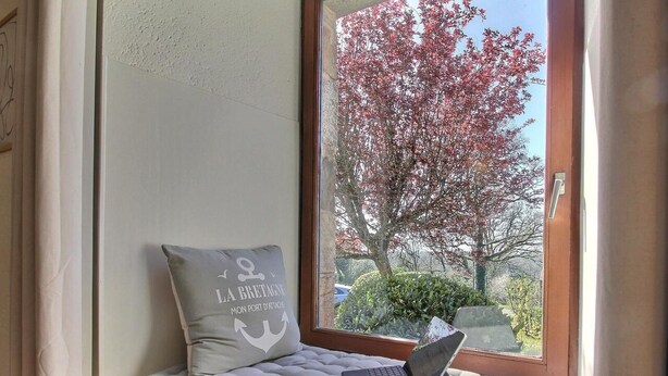Asiento junto a la ventana con una computadora portátil, un cojín con texto y vista a los árboles.