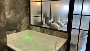 Ein Badezimmer mit einer großen Badewanne, einer Glasabtrennung und Blick auf ein Schlafzimmer mit Bett und Kissen.