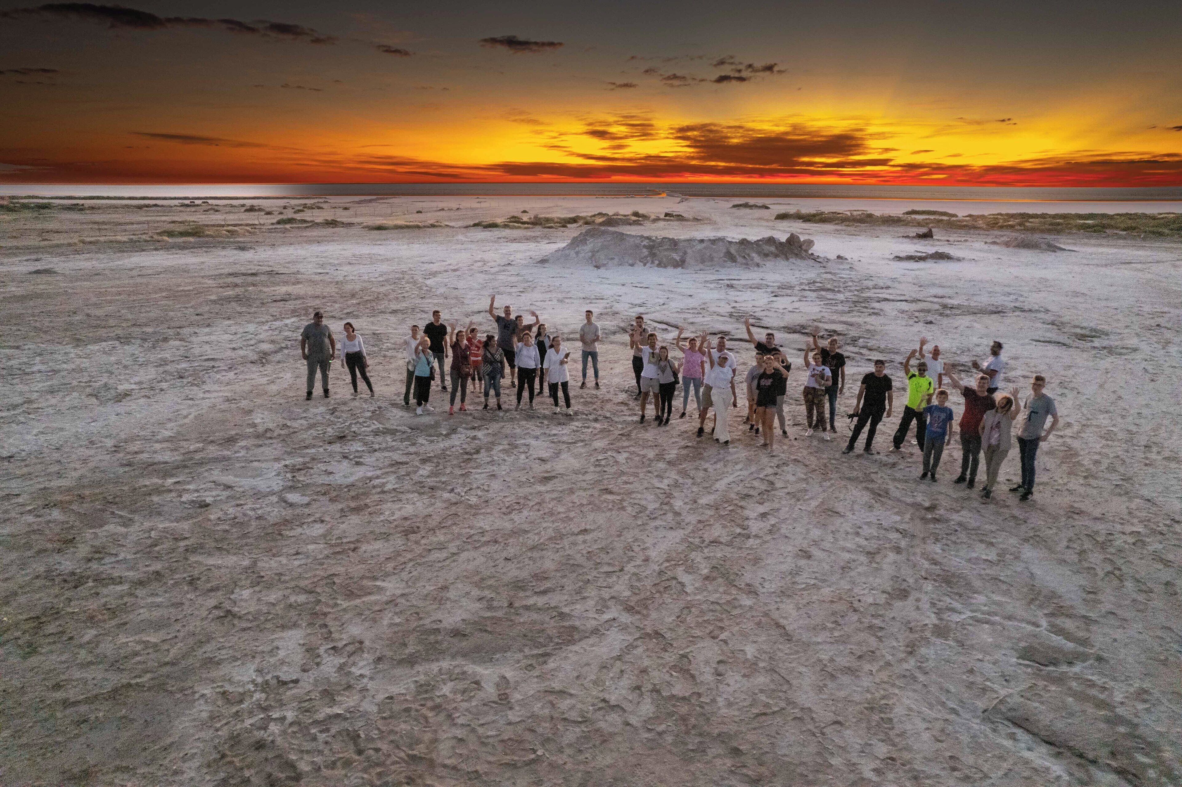 A group of people on a sandy beach with a sunset in the background.