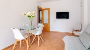 A modern living room with a glass dining table, white chairs, a grey sofa, a flat-screen TV mounted on the wall, and a wooden door. - Apartment 'Gran Playa Dreams' with Sea View, Wi-Fi and Air Conditioning (Santa Pola)