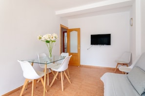 Un salon moderne avec une table à manger en verre, des chaises blanches, un canapé gris, un téléviseur à écran plat fixé au mur et une porte en bois.