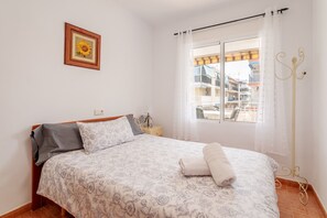 Una cama bien hecha con colcha de estampado floral, un cuadro enmarcados con un girasol en la pared, una ventana con vistas a edificios y un perchero de metal.