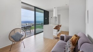Un salon moderne comprenant un canapé, un fauteuil, un petit coin bar et une grande porte-fenêtre coulissante donnant sur un balcon avec vue sur la mer.