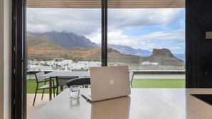 Une pièce avec un ordinateur portable posé sur une table, un verre d’eau et une vue sur des montagnes et une ville.