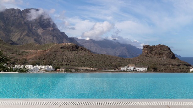 Une piscine d’un bleu éclatant, avec en arrière-plan des montagnes, des bâtiments blancs et un promontoire rocheux.
