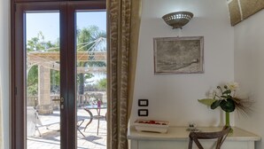 Una habitación con puerta de vidrio corrediza que da a un patio exterior, una pequeña mesa blanca con un florero y flores, un cuadro en la pared y una lámpara en el techo.