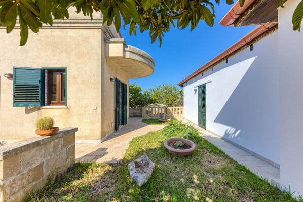 A courtyard with a stone wall, a potted cactus, a stone planter, and a pathway leading to a building with a curved corner and a window with shutters.