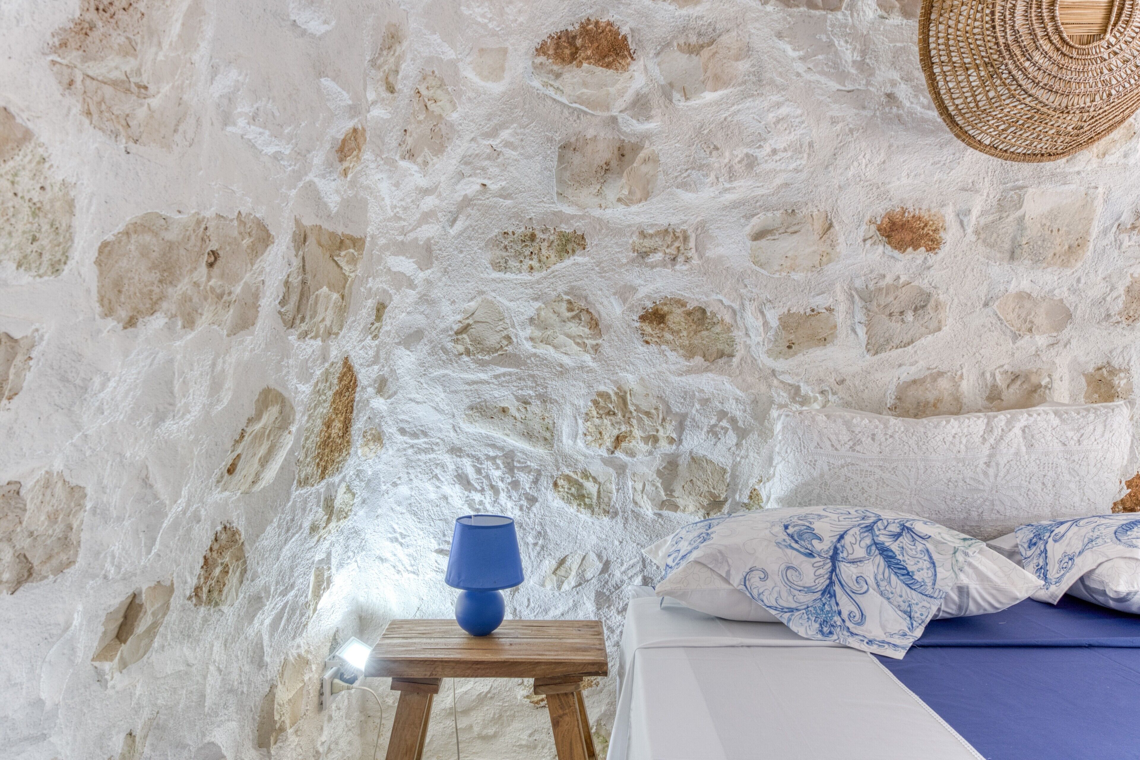 Une pièce avec un mur en pierre, une table de chevet en bois surmontée d’une lampe bleue, un lit avec une literie bleue et un panier en osier.
