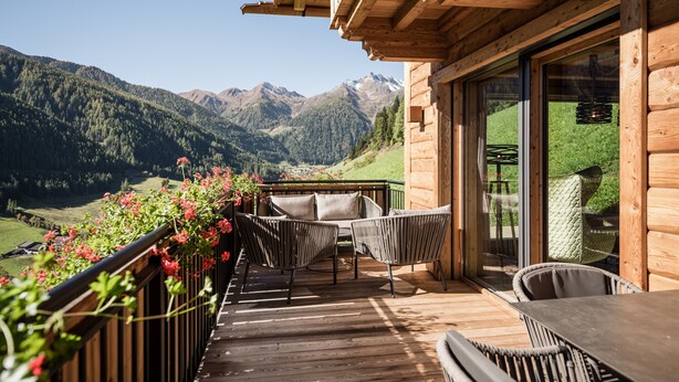 A wooden deck with wicker furniture, a glass door leading to a balcony, and a mountainous landscape in the background.