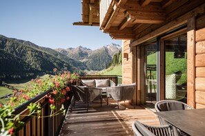 Una terraza de madera con mobiliario de mimbre, una puerta de cristal que da a un balcón y un paisaje montañoso al fondo.