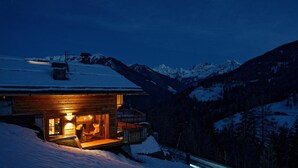 A wooden cabin with a porch and a ski chair, surrounded by snow-covered mountains.