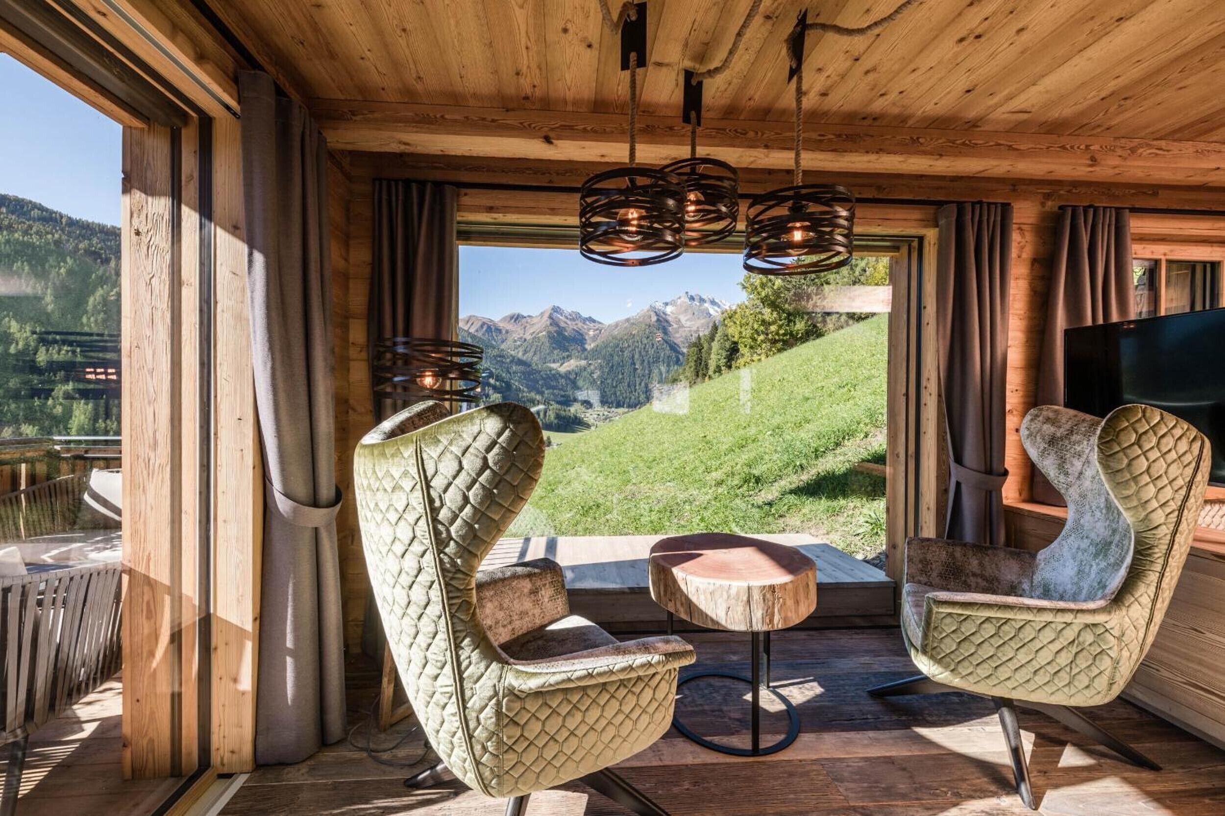 A wooden interior with a view of mountains, two upholstered chairs, a small round table, and a television.