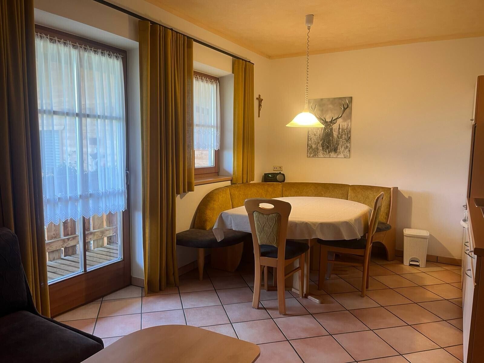 Un coin repas comprenant une table ronde, des chaises, un banc, une fenêtre avec des rideaux, une lampe fixée au mur et un tableau.