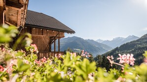 Una cabaña de madera con techo de tejas, rodeada de abundante vegetación y montañas al fondo.