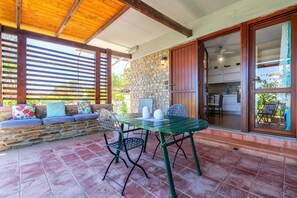 A covered patio with a stone wall, a wooden bench with cushions, a table with chairs, and a sliding glass door leading to a kitchen. - Holiday Home 'Casa 2 Punta Negra' with Sea View, Private Terrace and Air Conditioning (Stintino)