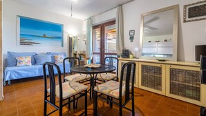 Sala de estar con sofá, mesa de comedor, sillas, ventilador de techo, un cuadro, un espejo y una zona de cocina con armarios.