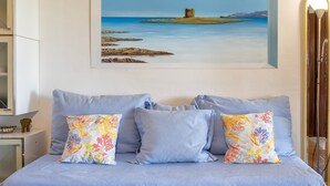 Una cama con ropa de cama azul claro, dos almohadas con motivos florales y un cuadro de un paisaje costero en la pared.