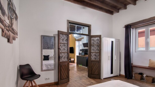 A bedroom with a bed, a chair, a wardrobe, and a view of the kitchen.