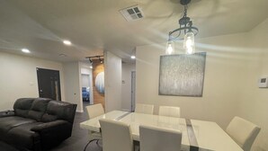 Una sala de estar moderna con un sofá de cuero, una mesa de comedor de vidrio con sillas blancas, iluminación empotrada en el techo y un elemento decorativo en la pared.