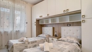 Ein Hotelzimmer mit zwei Betten, einem Holzkopfende, einem Nachttisch und einem Fenster mit durchscheinenden Vorhängen.