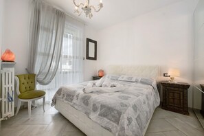 Une chambre à coucher comprenant un lit, une chaise, une table de chevet, un radiateur et une fenêtre avec des rideaux.