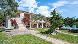 A two-story building with a stone staircase leading to a balcony, surrounded by greenery and a clear sky.