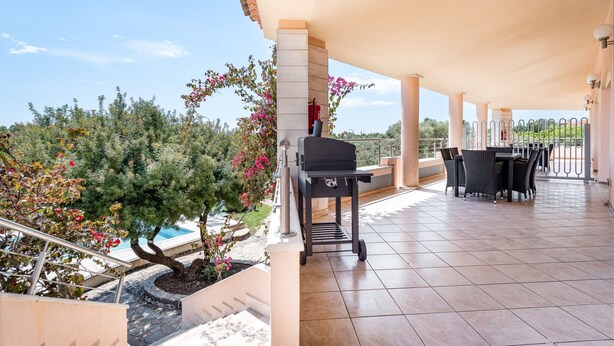 A covered patio area with a grill, outdoor seating, and a view of the pool and garden.