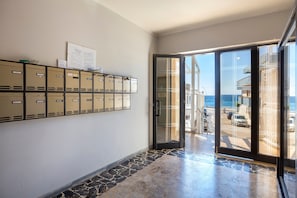 Un pasillo con taquillas, una puerta de vidrio que da a un balcón con vistas al mar, y suelo de baldosas.