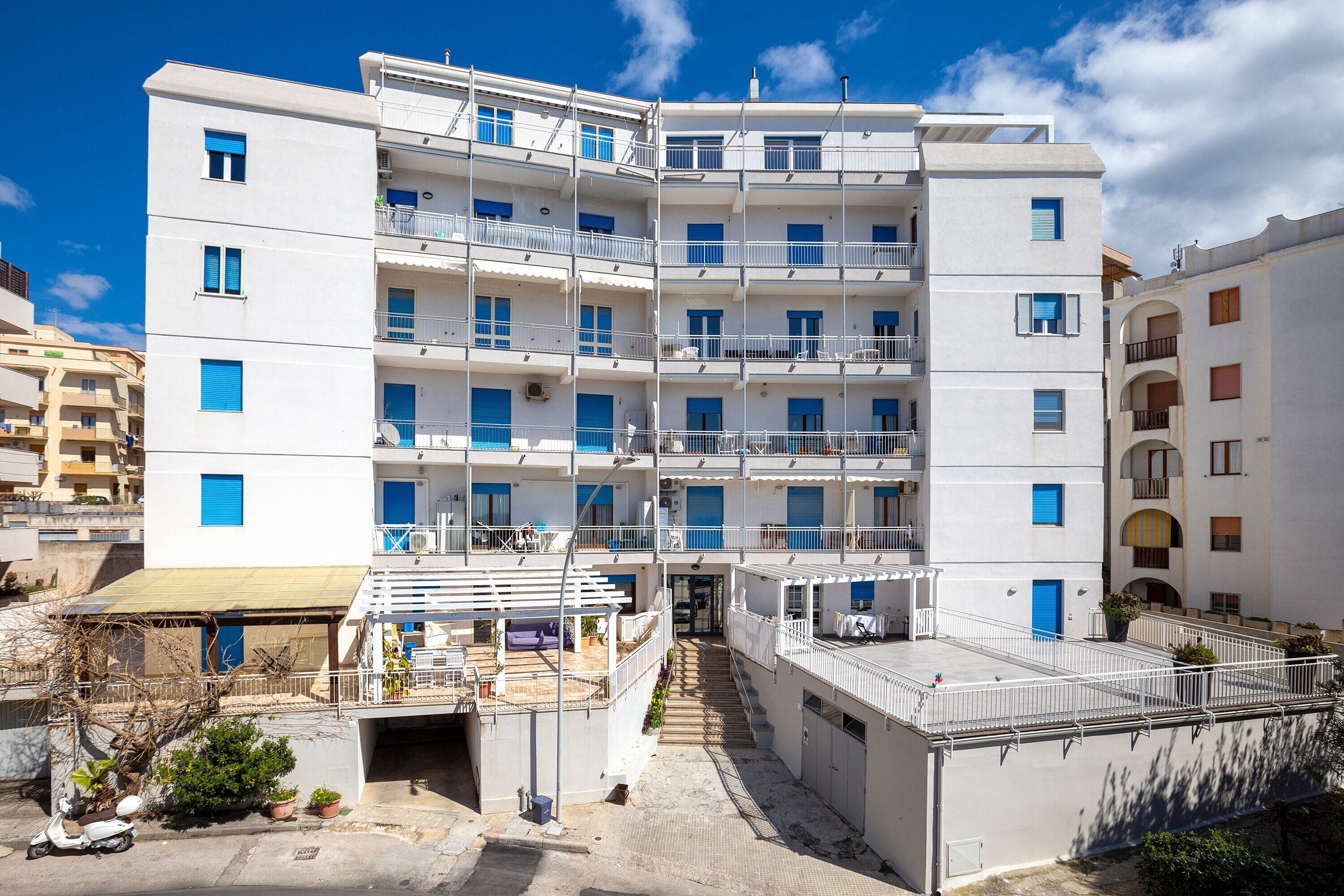 A multi-story residential building with balconies, a covered outdoor area, and a staircase leading to the entrance.