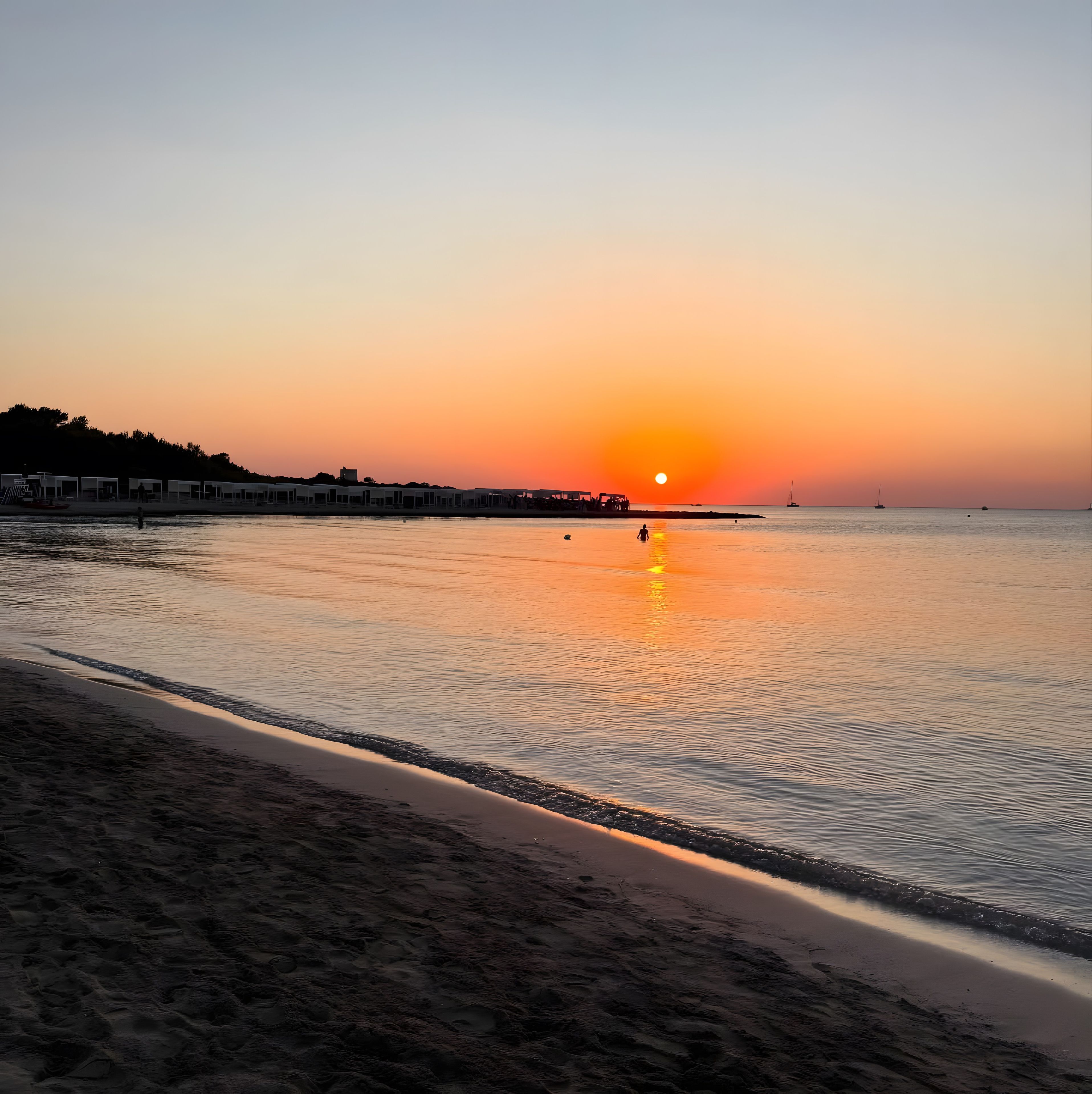Una playa con un atardecer, agua tranquila y una hilera de edificios a lo largo de la costa.