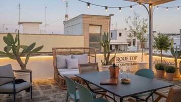 An outdoor seating area with a table, chairs, and a cushioned bench. There are potted plants and a sign that reads 'ROOF TOP SANPIETRAN.'