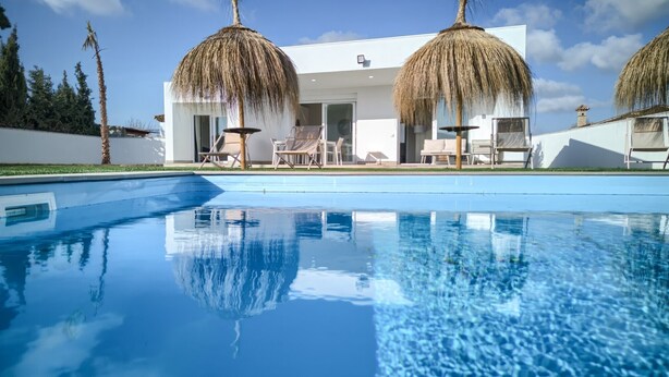 A modern pool with clear blue water, a white building with large windows, and two thatched umbrellas. - Villa 'Sanctipetri 10 Pax – adults over 25 only' with private pool, Wi-Fi & air conditioning (Chiclana de la Frontera)