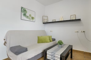 Un salon comprenant un canapé, des coussins, une petite table avec une plante, une étagère murale ornée d’objets décoratifs et un tableau encadré.