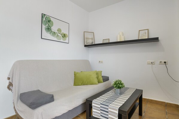 A living room with a sofa, pillows, a small table with a plant, a wall-mounted shelf with decorative items, and a framed picture. - Apartment 'Antonia Conil' with Shared Terrace, Wi-Fi and Air Conditioning (Conil de la Fra.)
