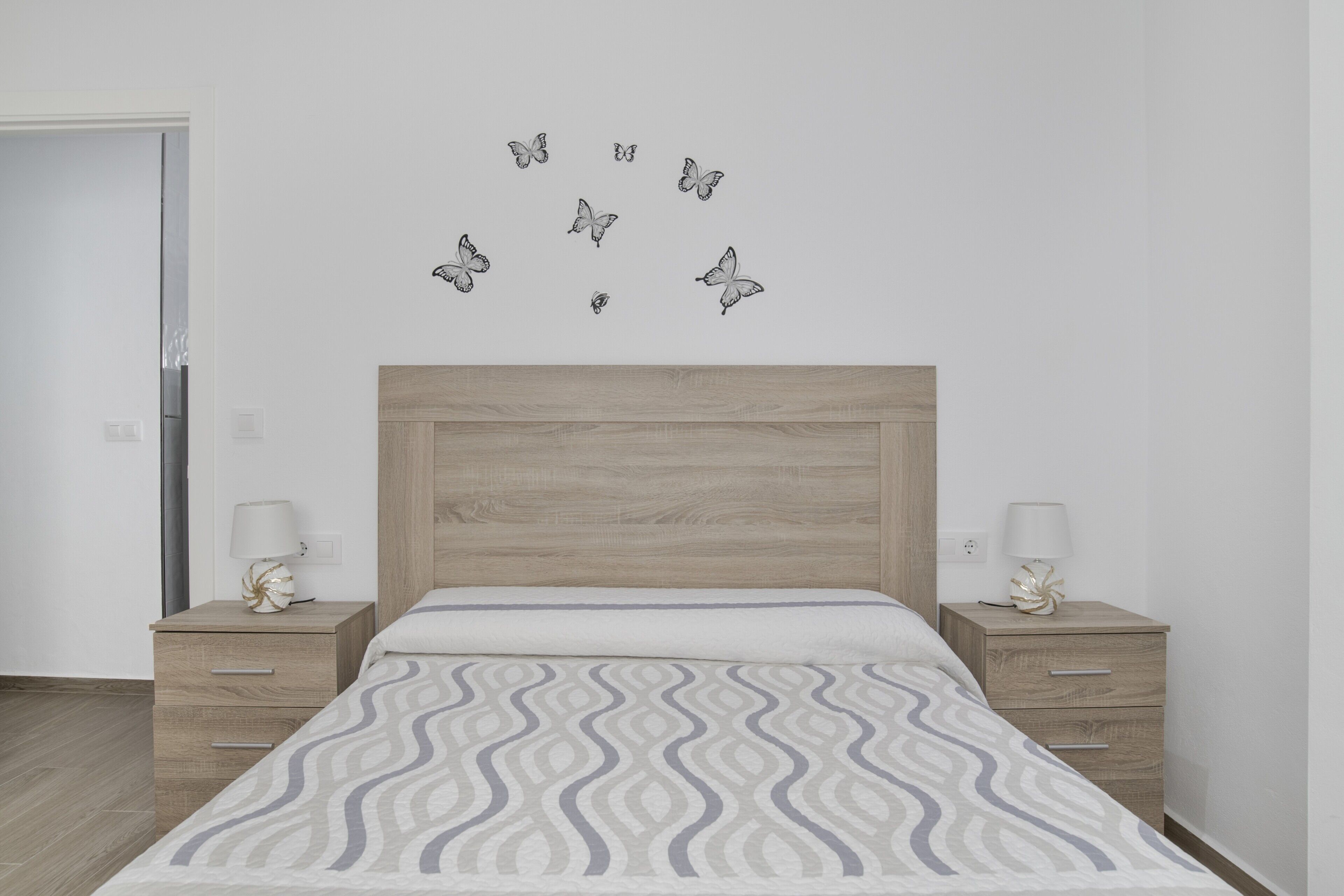 Een slaapkamer met een houten hoofdbord, een bed met een decoratieve spreidek en twee nachtkastjes met lampen, plus wanddecoratie van vlinders.