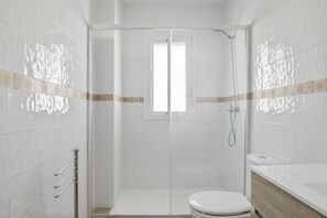 A bathroom with white tiles, a shower, a toilet, and a window.