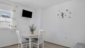 Ein Essbereich mit einem weißen Tisch und Stühlen, einer Topfpflanze, einem an der Wand befestigten Fernseher und einer Wandsticker-Darstellung einer tanzenden Person mit Schmetterlingen.