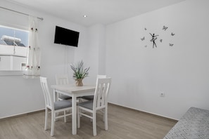 Un coin repas avec une table et des chaises blanches, une plante en pot, un téléviseur fixé au mur et un motif décoratif représentant une silhouette dansante entourée de papillons.