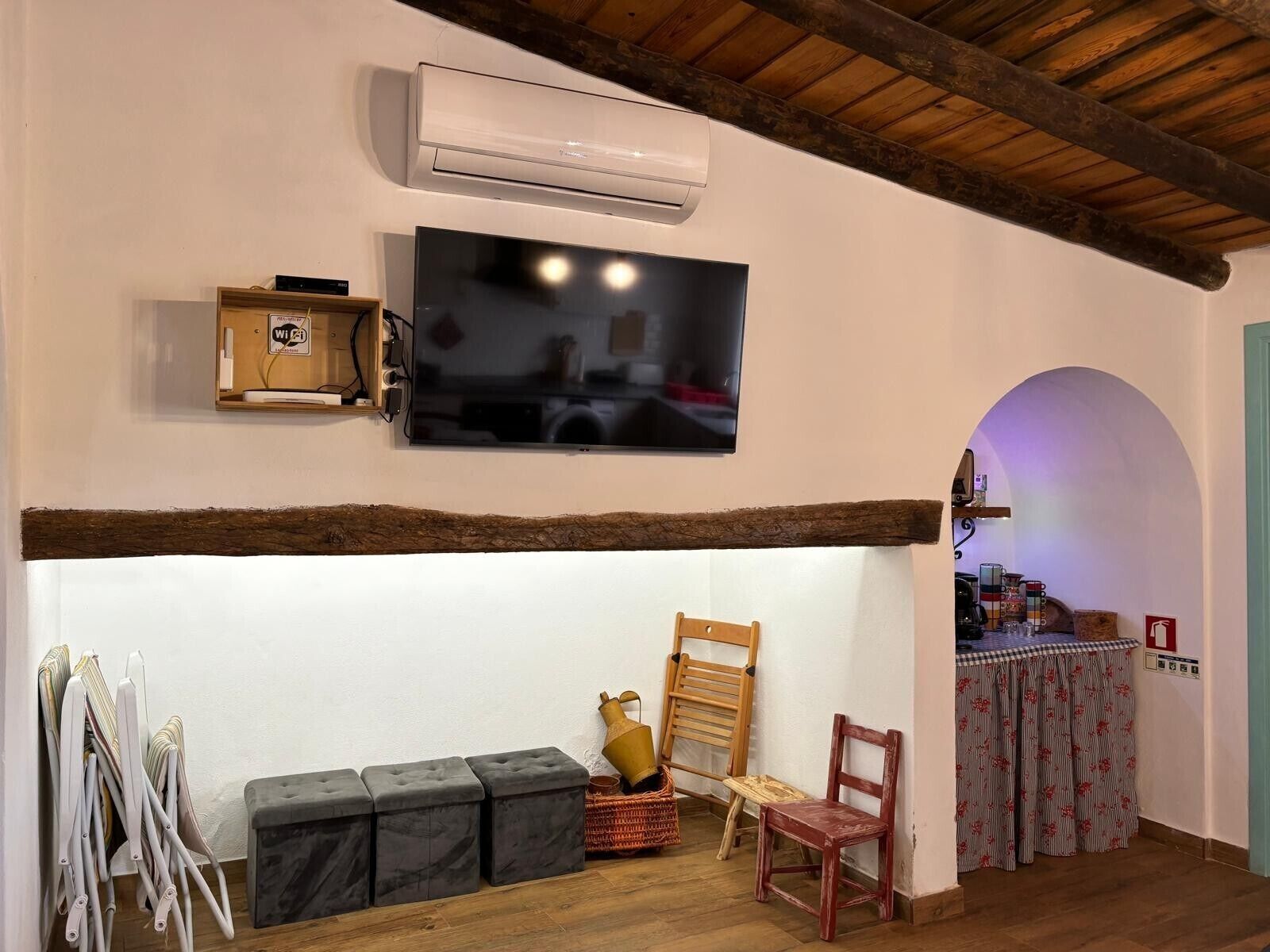 Una habitación con una televisión montada en la pared, techo de madera, un banco, una silla y una mesa con mantel floreado.